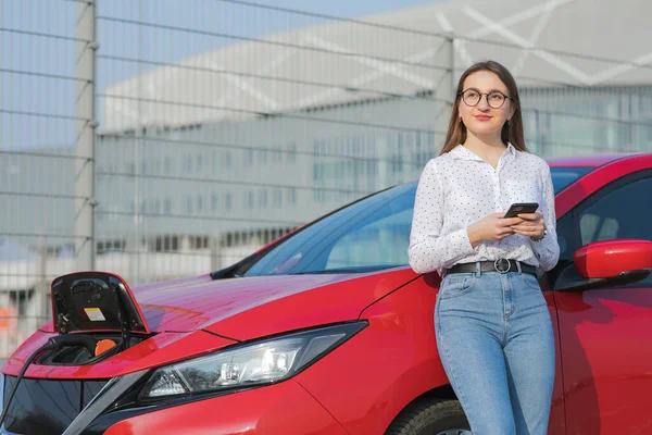 Vit flicka med hjälp av smart telefon och väntande strömförsörjning ansluta till elfordon för att ladda batteriet i bilen. Ekologisk bil ansluten och ladda batterier. — Stockfoto