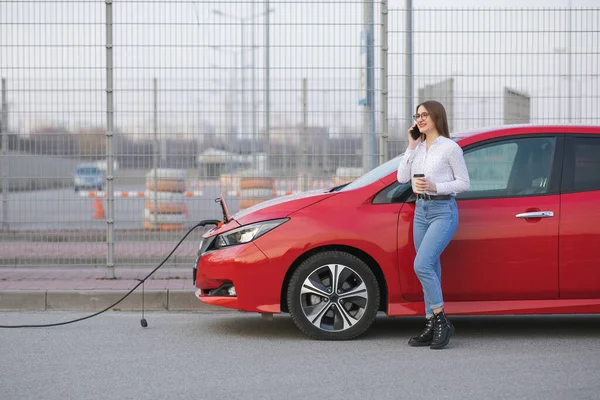 Elektrikli araba caddede şarj oluyor. Ekolojik Araba Bağlı ve Şarj Bataryaları. Akıllı Telefon ve Bekleme Güç Aracını Şarj Etmek İçin Elektrikli Araçlara Bağlanırken Kahve İçeceği Kullan
