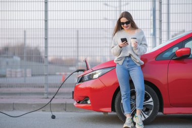 Elektrikli araba caddede şarj oluyor. Ekolojik Araba Bağlı ve Şarj Bataryaları. Akıllı Telefon ve Bekleme Güç Aracını Şarj Etmek İçin Elektrikli Araçlara Bağlanırken Kahve İçeceği Kullan