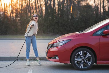 Elinde elektrik kablosu tutan mutlu genç kız. Elektrikli otomobilin yanında duruyor ve kameraya bakıyor..
