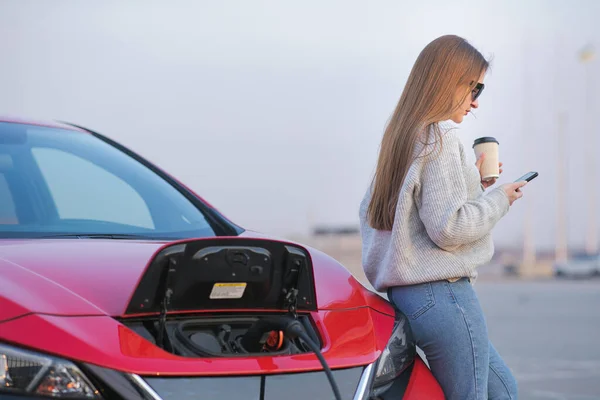 Flicka använda kaffe dricka medan du använder Smart telefon och väntar strömförsörjning Anslut till elektriska fordon för laddning av batteriet i bilen. — Stockfoto