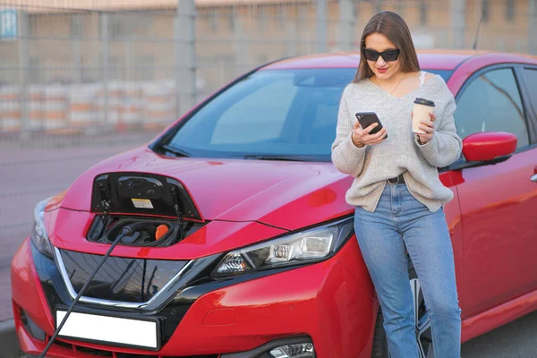 Žena stojí s telefonem u svého elektrického auta a čeká, až se vozidlo nabije. Nabíjení elektromobilu — Stock fotografie