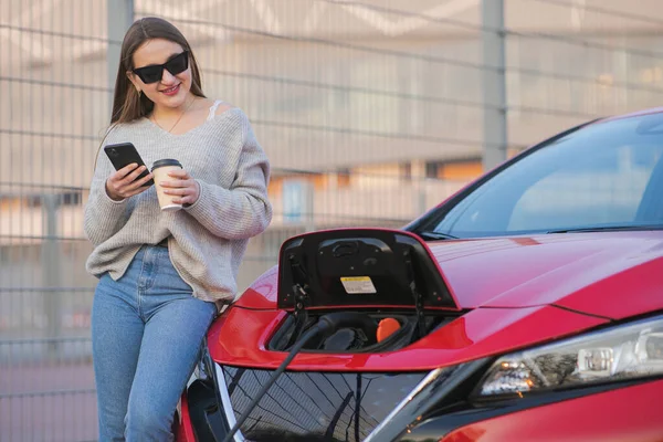 Elektrické auto nabíjí na ulici. Ekologické auto připojené a nabíjení baterie. Girl Use Coffee Drink While Using SmartPhone and Waiting Power Supply Connect to Electric Vehicles for Charging — Stock fotografie