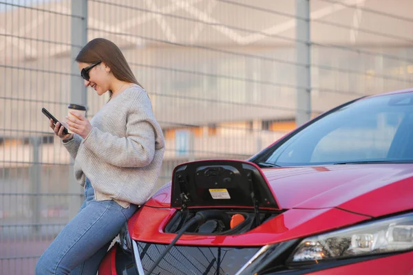 Elektrické auto nabíjí na ulici. Ekologické auto připojené a nabíjení baterie. Girl Use Coffee Drink While Using SmartPhone and Waiting Power Supply Connect to Electric Vehicles for Charging — Stock fotografie