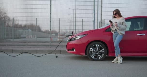 Fata elegantă stă cu telefonul lângă mașina ei electrică roșie și așteaptă când vehiculul se va încărca. Conectarea încărcătorului unui automobil electric . — Videoclip de stoc