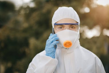 Koruyucu PPE takımlı kendine güvenen Asyalı doktor telefonda konuşuyor. Kimyasal korumalı kız telefonda konuşuyor. Doktor epidemiyoloji uzmanı Coronavirus COVID-19 ile savaşıyor..