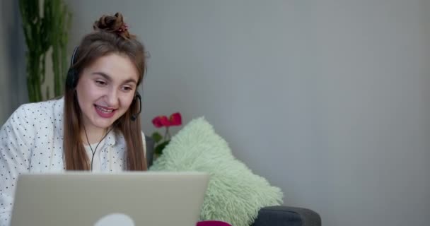 Glad flicka e-lärande med bärbar dator och hörlurar sitter på en soffa i vardagsrummet hemma — Stockvideo