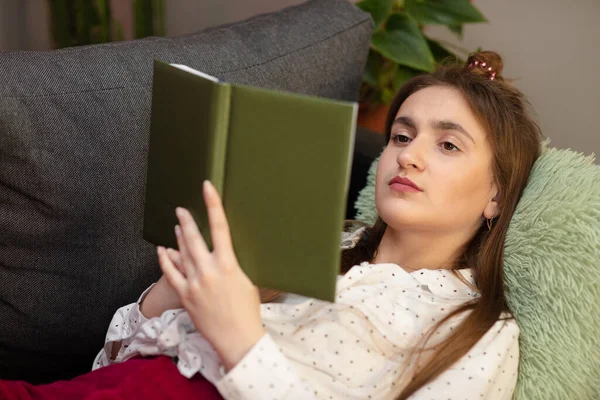 Portret uśmiechniętej kobiety trzymającej książkę i leżącej na kanapie. Nastolatka w domu - student czytać książki leżące na kanapie — Zdjęcie stockowe