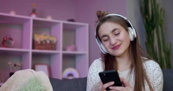 Flicka lyssnar på musik genom hörlurar och använda smartphone koppla av på soffan hemma ha kul. Prylar, glad ungdom och fritid koncept — Stockvideo