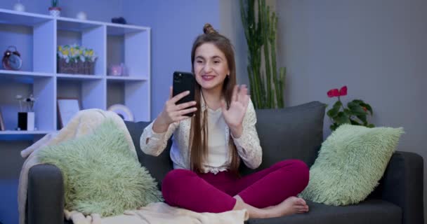 現代のスマート電話波手こんにちはを保持幸せな若い女性ブロガーのインフルエンサー。笑顔vlogger女の子見ますでモバイル作るビデオ呼び出し,撮影vlog取ります自画撮り孤立した上のホーム背景 — ストック動画