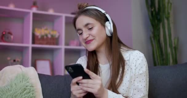 Χαρούμενη κοπέλα στα ακουστικά είναι ο χορός στον καναπέ και τη χρήση smartphone αφής οθόνη. Σύγχρονη έννοια τεχνολογίας — Αρχείο Βίντεο