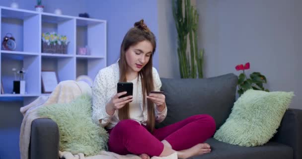 Молода жінка купує онлайн з кредитною карткою, сидячи на дивані у вітальні. Жінка використовує смартфон і здійснює онлайн транзакції — стокове відео