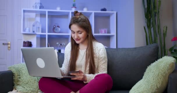 Mädchen verbindet sich mit einem Laptop und erledigt Online-Einkäufe mit einer Kreditkarte. Junge Mädchen kaufen online mit Kreditkarte, während sie auf dem Sofa sitzen. Frau benutzt Laptop und erledigt Online-Transaktionen. — Stockvideo