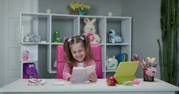 Ritratto di una bambina in età prescolare che guarda il libro e sorride. Autoisolamento e scolarizzazione online. Apprendimento a distanza, scuola online . — Video Stock
