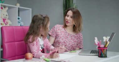 Kadın öğretmen ya da koruyucu anne mutfak masasında oturan beyaz kız öğrencilere yardım ediyor. Çeşitli dadılar ve çocuklar evde ders çalışarak not defterinde yazmayı öğreniyorlar.