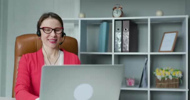 Ο διαχειριστής εξυπηρέτησης πελατών μιλάει στην κάμερα που επικοινωνεί εξ αποστάσεως στο γραφείο του σπιτιού. Γυναίκα υπάλληλος γραφείου με αυτοπεποίθηση φοράει ακουστικά, τηλεδιάσκεψη καλώντας, κοιτάζοντας το laptop, υπολογιστή — Αρχείο Βίντεο