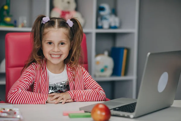 Gülümseyen sevimli ilkokul çocuğu öğrenme sınıfında tek başına masada dizüstü bilgisayarla oturuyor. Güzel kız ödevini yapıyor. — Stok fotoğraf
