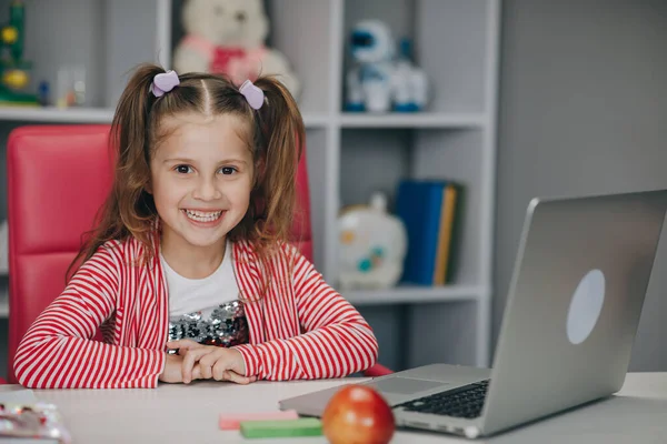 Portret van een kleuter die naar de camera kijkt en glimlacht. Kind meisje zit aan het bureau in de kinderkamer thuis en studeert online. afstandsonderwijs, school online — Stockfoto