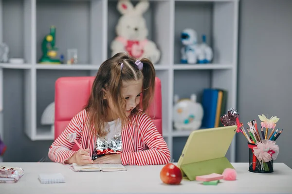 Anaokulundaki kız çevrimiçi ders izliyor ve evden ders çalışıyor. Bilgisayar ekranına bakarken not alan bir kız çocuğu profesörün video görüşmesinde matematik yapmasını izliyor. — Stok fotoğraf