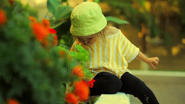 Klein meisje in een gestreepte T-Shirt groen hoed en zwarte legging zittend op de rand van een tuin Bed en werken op de grond — Stockvideo