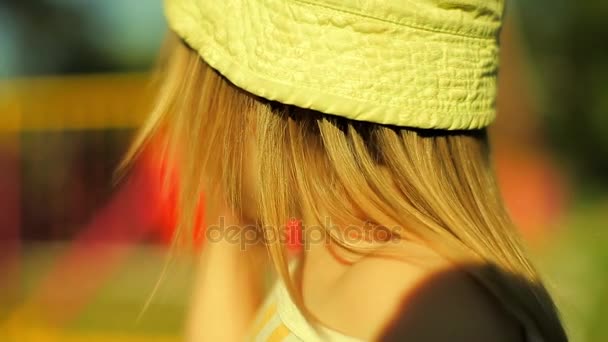 Kleines Mädchen in grünem Panama mit goldenem Haar nahm Löwenzahn in den Mund und schaute sich das Schaukelkarussell im Park an — Stockvideo