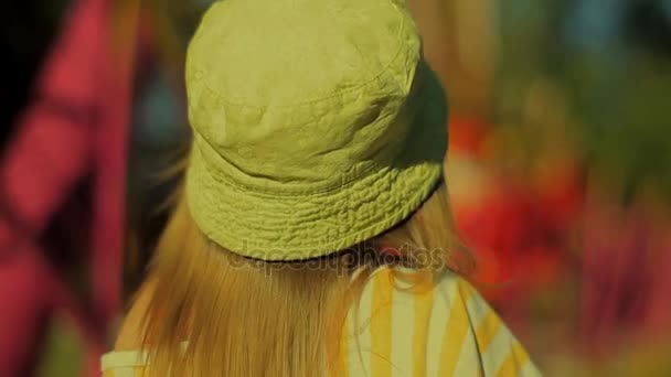 Utsikten från den tillbaka bedårande tjejen med blont hår i en grön hatt tittar på svängande Carousel och sedan vänder och går bort — Stockvideo