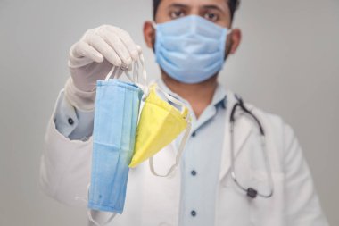 Roman Coronavirus COVID-19 - Beyaz önlüklü doktor, beyaz eldivenli doktor elinde beyaz arka planda tekstil tıbbi maskeleri, korona virüslerine ve bakterilere karşı koruyucu aksesuarlar tutuyor.