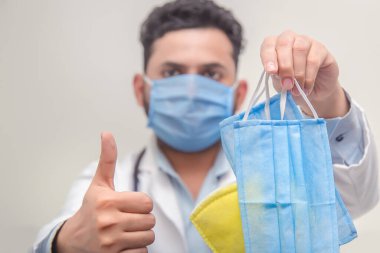 Beyaz önlüklü doktor, beyaz eldivenli doktor beyaz arka planda tekstil tıbbi maskeleri tutuyor. Diğer elinde ise korona virüslerine ve bakterilere karşı koruyucu aksesuarlar var.