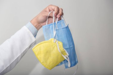 Beyaz önlüklü doktor, elinde beyaz arka planda tekstil tıbbi maskeleri, diğer elinde de maskeyi, korona virüslerine ve bakterilere karşı koruyucu aksesuar tutuyor.