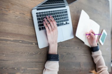 Bilgisayarlı, tabletli bir ofis masası. Kadın ellerinde fuşya renkli bir kalem. Yazmak için sarı çarşaflar, kalemler ve masanın üstündeki saksıda bir çiçek.