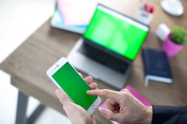 Bilgisayarı, tableti ve telefonu olan bir ofis masası. Adamın elleri yeşil ekranlı bir akıllı telefonu tutuyor. Ofis İşçi Masası