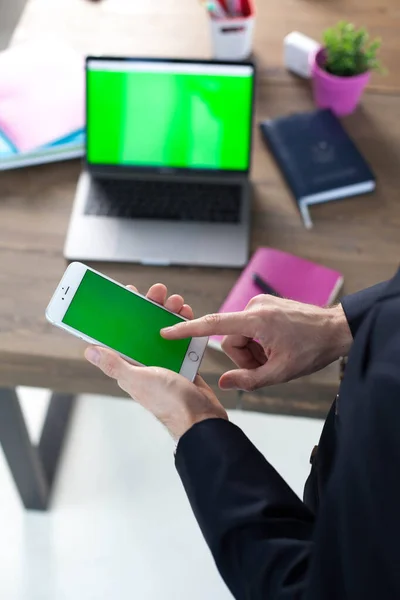 Office desk with laptop, tablet and phone. Man\'s hands are holding a smartphone with a green screen. Office Worker Desk