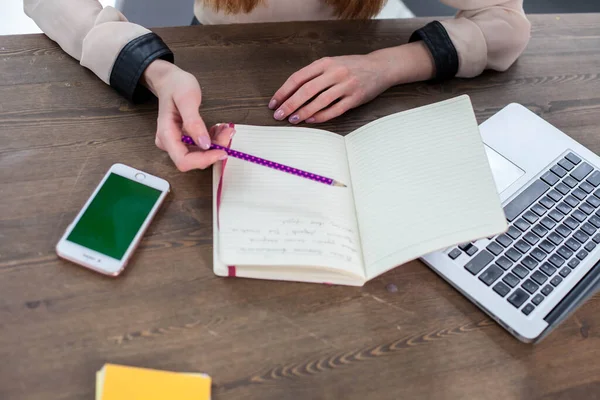 Office desk with laptop, tablet. In female hands a fuchsia-colored pencil. Yellow sheets for writing, pencils and a flower in a pot on the table