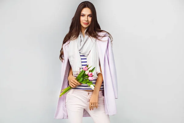 Foto de moda de una hermosa joven con tulipanes en la mano .she vestido con un hermoso abrigo, bufanda, pantalones y camiseta con rayas — Foto de Stock