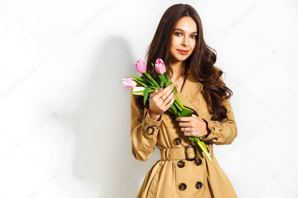 Fashion photo of a beautiful young woman with tulips in her hand .she dressed in a beautiful brown trench coat, crimson dress.Spring concept. March 8. beautiful girl in stylish clothes.