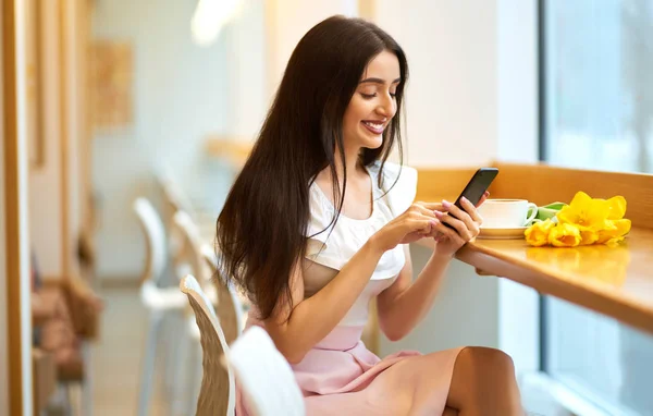 Beautiful Young Woman With Spring Tulips Flowers Bouquet At Cafe. Woman Drinking Coffee And Using Phone. Spring Style. Brunette Woman In Nice Spring.Beautiful Spring Mood. Fashion Spring/Summer.