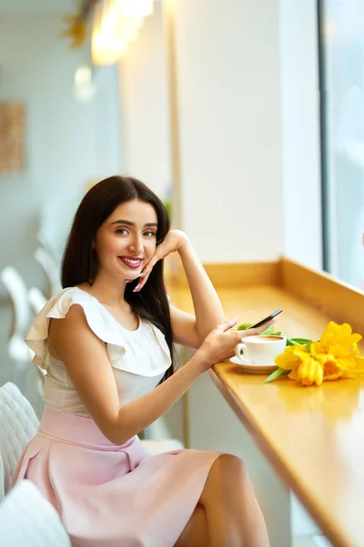 Beautiful Young Woman With Spring Tulips Flowers Bouquet At Cafe. Woman Drinking Coffee And Using Phone. Spring Style. Brunette Woman In Nice Spring.Beautiful Spring Mood. Fashion Spring/Summer.