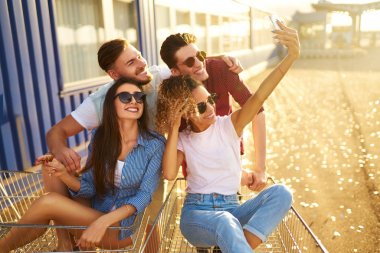 Selfie Zamanı! Grup alışveriş arabaları üzerinde eğleniyor mutlu genç insan. Alışveriş sepeti yarış çok ırklı çocuklar. Güzel yaz günü güneş ışığı ile. Yaşam tarzı kavramı 
