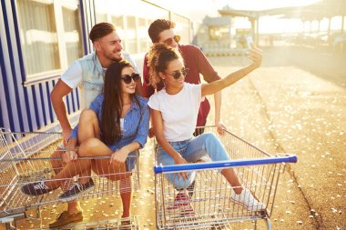 Selfie Zamanı! Grup alışveriş arabaları üzerinde eğleniyor mutlu genç insan. Alışveriş sepeti yarış çok ırklı çocuklar. Güzel yaz günü güneş ışığı ile. Yaşam tarzı kavramı 