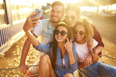 Selfie Zamanı! Grup alışveriş arabaları üzerinde eğleniyor mutlu genç insan. Alışveriş sepeti yarış çok ırklı çocuklar. Güzel yaz günü güneş ışığı ile. Yaşam tarzı kavramı 