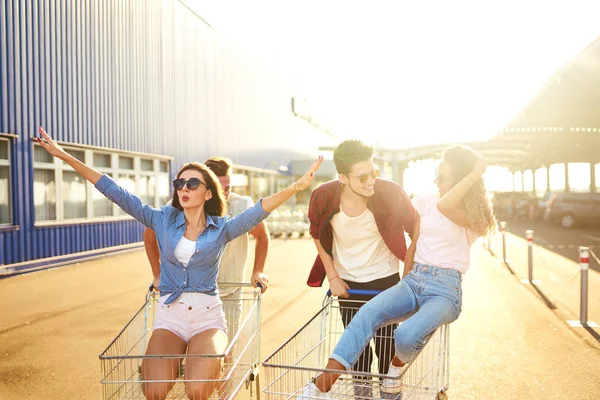 Group of happy young people having fun on shopping trolleys. Multiethnic young people racing on shopping cart. Beautiful summer day with sunlight. Lifestyle concept. Group of friends enjoy life.