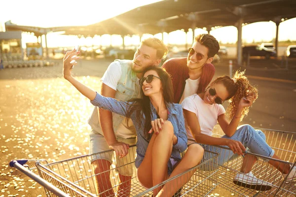 Selfie Zamanı! Grup alışveriş arabaları üzerinde eğleniyor mutlu genç insan. Alışveriş sepeti yarış çok ırklı çocuklar. Güzel yaz günü güneş ışığı ile. Yaşam tarzı kavramı 