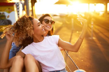 Selfie Zamanı! İki güzel kız alışveriş arabaları üzerinde eğleniyor. Alışveriş sepeti yarış çok ırklı çocuklar. Güzel yaz günü güneş ışığı ile. Yaşam tarzı kavramı.