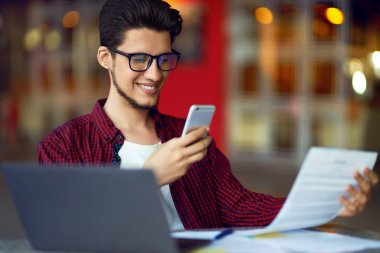 Masanın üstünde laptop ile gözlük gülümseyen genç hipster adam. Programcı, web geliştirici, mobil ve Masaüstü Web sürümleri karşılaştırarak ofiste çalışan tasarımcı.