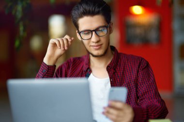 Masanın üstünde laptop ile gözlük gülümseyen genç hipster adam. Programcı, web geliştirici, mobil ve Masaüstü Web sürümleri karşılaştırarak ofiste çalışan tasarımcı.