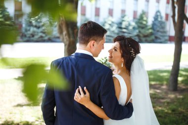 Gelin ve damat parkta birlikte yürürler. Beyaz elbiseli çekici gelin, damat koyu renkli şık bir takım elbise giyiyor. Güzel gelin ve şık damat. Düğün günü. Evlilik..