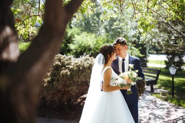 Gelin ve damat parkta birlikte yürürler. Beyaz elbiseli çekici gelin, damat koyu renkli şık bir takım elbise giyiyor. Güzel gelin ve şık damat. Düğün günü. Evlilik..