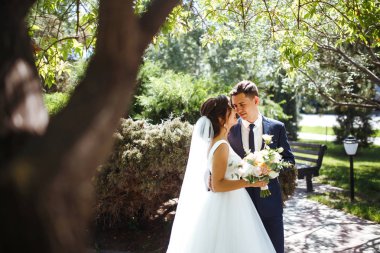 Gelin ve damat parkta birlikte yürürler. Beyaz elbiseli çekici gelin, damat koyu renkli şık bir takım elbise giyiyor. Güzel gelin ve şık damat. Düğün günü. Evlilik..