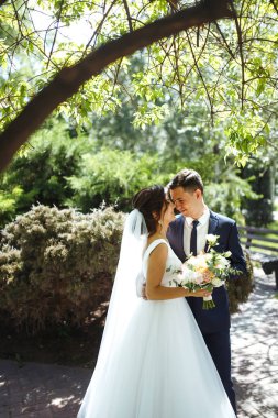 Gelin ve damat parkta birlikte yürürler. Beyaz elbiseli çekici gelin, damat koyu renkli şık bir takım elbise giyiyor. Güzel gelin ve şık damat. Düğün günü. Evlilik..