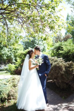 Gelin ve damat parkta birlikte yürürler. Beyaz elbiseli çekici gelin, damat koyu renkli şık bir takım elbise giyiyor. Güzel gelin ve şık damat. Düğün günü. Evlilik..